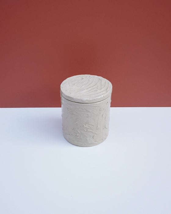 A small cylindrical lidded container with a textured, hand-painted finish in a light warm gray-beige color, set against a split background of white and terracotta red. Brutes Ceramics | Hope Collection | Shop Fodder