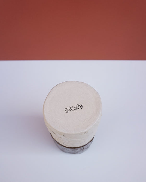 A handmade ceramic tin with a round, flat lid, featuring a stamped 'brutes' logo on the top. The tin has a textured, hand-painted beige finish with a dark brown base, set against a split white and terracotta background. Brutes Ceramics | Hope Collection | Shop Fodder