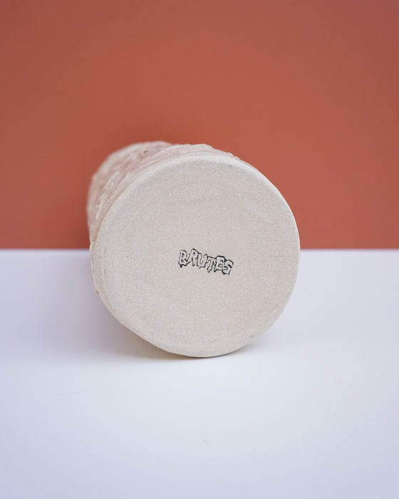 A cylindrical ceramic vase or planter shown from its bottom, featuring a textured beige body and a stamped black brand logo that reads 'brutes'. The vase is positioned on a white surface against a terracotta-colored background.  Brutes Ceramics | Hope Collection | Shop Fodder