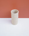 A light beige textured ceramic cylinder vase with a ribbed, organic pattern, standing on a white surface against a terracotta-colored background. Brutes Ceramics | Hope Collection | Shop Fodder