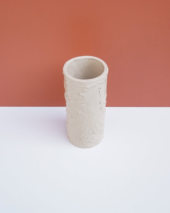 A light beige textured ceramic cylinder vase with a ribbed, organic pattern, standing on a white surface against a terracotta-colored background. Brutes Ceramics | Hope Collection | Shop Fodder
