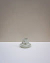 A small light green ceramic candle holder with a matching saucer, centered on a white surface against a warm beige background. Emma Johnson Ceramics | Fodder | Hope Collection