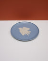 A round blue ceramic plate with a textured beige bird design in the center, set against a split white and terracotta background. Fern Taylor Ceramics | Hope Collection | Shop Fodder