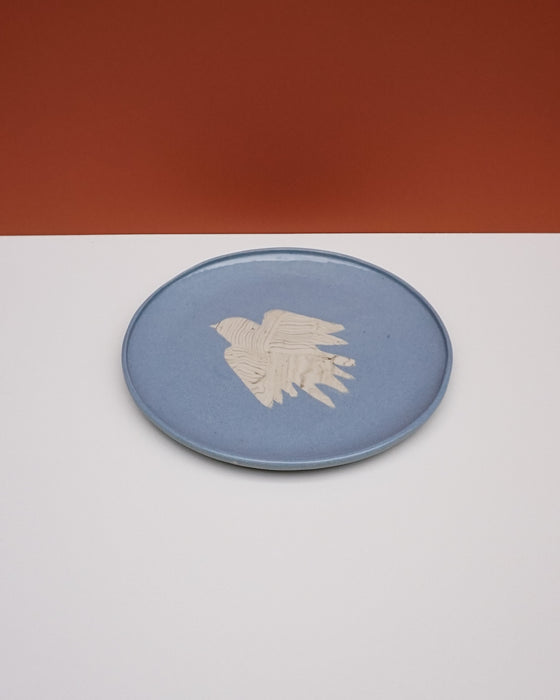 A round blue ceramic plate with a textured beige bird design in the center, set against a split white and terracotta background. Fern Taylor Ceramics | Hope Collection | Shop Fodder