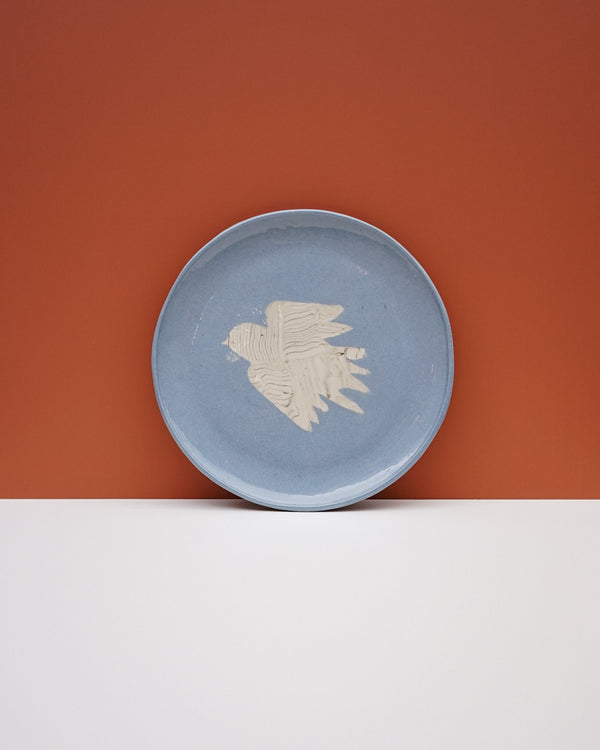 A round light blue ceramic plate with a textured off-white bird design in the center, set against a split orange and white background. Fern Taylor Ceramics | Hope Collection | Shop Fodder