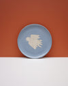 A round light blue ceramic plate with a textured off-white bird design in the center, set against a split orange and white background. Fern Taylor Ceramics | Hope Collection | Shop Fodder