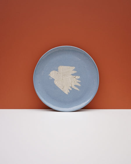 A round light blue ceramic plate with a textured off-white bird design in the center, set against a split orange and white background. Fern Taylor Ceramics | Hope Collection | Shop Fodder