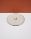 A round ceramic plate with a warm cream glaze and a small blue floral motif in the center, set against a split orange and white background. Fern Taylor Ceramics | Hope Collection | Shop Fodder