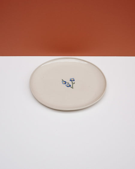 A round ceramic plate with a warm cream glaze and a small blue floral motif in the center, set against a split orange and white background. Fern Taylor Ceramics | Hope Collection | Shop Fodder
