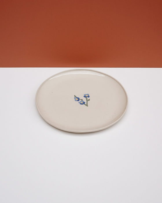 A round ceramic plate with a warm cream glaze and a small blue floral motif in the center, set against a split orange and white background. Fern Taylor Ceramics | Hope Collection | Shop Fodder