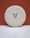 A round, off-white ceramic plate with a small blue and green floral design in the center, set against a split background of terracotta and white. Fern Taylor Ceramics | Hope Collection | Shop Fodder