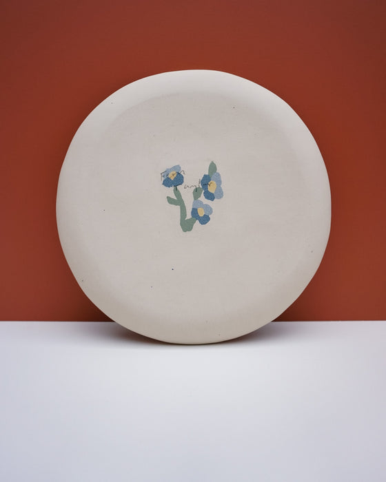 A round, off-white ceramic plate with a small blue and green floral design in the center, set against a split background of terracotta and white. Fern Taylor Ceramics | Hope Collection | Shop Fodder