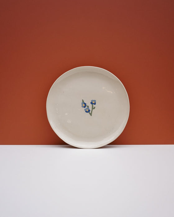 A round off-white ceramic plate with a small blue and green floral motif in the center, set against a split orange and white background. Fern Taylor Ceramics | Hope Collection | Shop Fodder