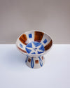 A handmade ceramic pedestal bowl with a white base, decorated with blue and brown geometric patterns, set against a neutral gray and white background.  Hoodmor Studio | Fodder | Hope Collection