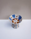 A handmade ceramic pedestal bowl with a white base, decorated with blue painted stripes, brown geometric accents, and a white star motif on the inside, set against a neutral gray and white background. Hoodmor Studio | Fodder | Hope Collection