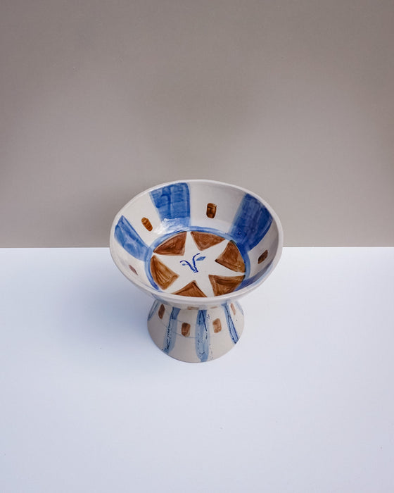 A handmade ceramic pedestal bowl with a white base, decorated with blue painted stripes, brown geometric accents, and a white star motif on the inside, set against a neutral gray and white background. Hoodmor Studio | Fodder | Hope Collection