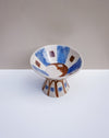 A handmade ceramic pedestal bowl with a wide rim and pedestal base, decorated with blue brushstrokes, brown geometric shapes, and a central brown crescent moon motif on a white background. Hoodmor Studio | Fodder | Hope Collection
