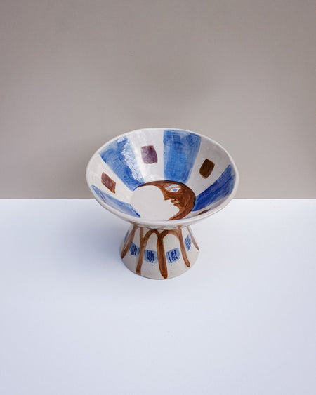 A handmade ceramic pedestal bowl with a wide rim and pedestal base, decorated with blue brushstrokes, brown geometric shapes, and a central brown crescent moon motif on a white background. Hoodmor Studio | Fodder | Hope Collection