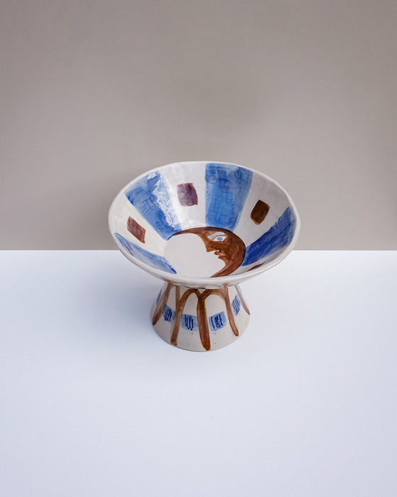 A handmade ceramic pedestal bowl with a wide rim and pedestal base, decorated with blue brushstrokes, brown geometric shapes, and a central brown crescent moon motif on a white background. Hoodmor Studio | Fodder | Hope Collection