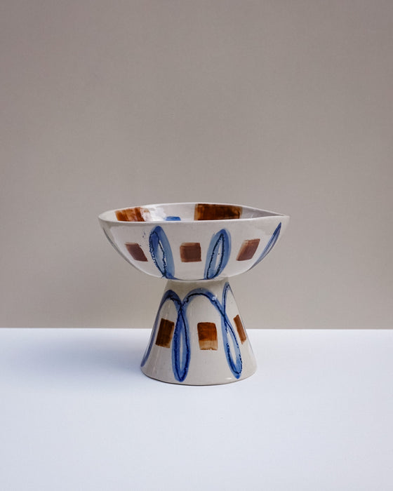 A ceramic pedestal bowl with a geometric pattern of blue ovals and brown squares on a white base, set against a neutral beige and white  Hoodmor Studio | Fodder | Hope Collectionbackground.