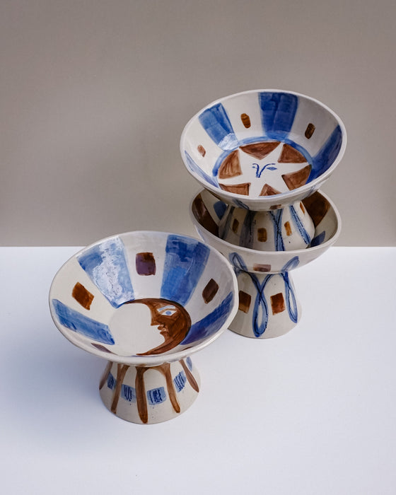 Three hand-painted ceramic high fruit bowls with a Mediterranean style, featuring blue and brown patterns on a white background, arranged on a neutral studio surface. Hoodmor Studio | Fodder | Hope Collection
