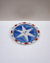 An oval ceramic plate with a hand-painted blue and white star design, featuring a red and blue patterned rim, set against a split white and gray background. Hoodmor Studio | Fodder | Hope Collection