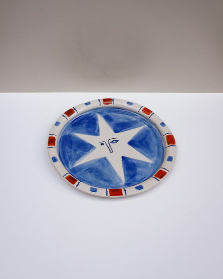 An oval ceramic plate with a hand-painted blue and white star design, featuring a red and blue patterned rim, set against a split white and gray background. Hoodmor Studio | Fodder | Hope Collection