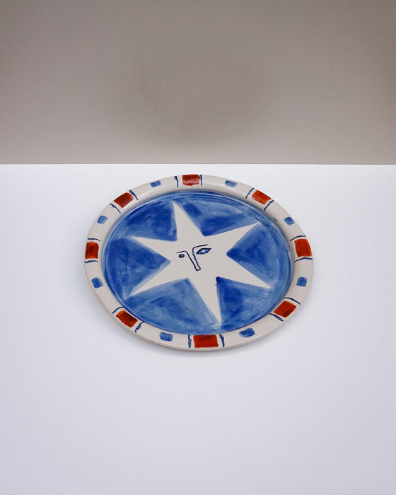 An oval ceramic plate with a hand-painted blue and white star design, featuring a red and blue patterned rim, set against a split white and gray background. Hoodmor Studio | Fodder | Hope Collection