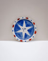 A round ceramic plate with a hand-painted blue and white star design, featuring a patterned rim with red and blue accents, set against a neutral gray and white background. Hoodmor Studio | Fodder | Hope Collection