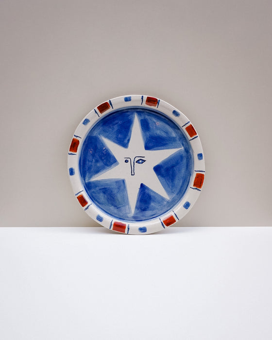 A round ceramic plate with a hand-painted blue and white star design, featuring a patterned rim with red and blue accents, set against a neutral gray and white background. Hoodmor Studio | Fodder | Hope Collection