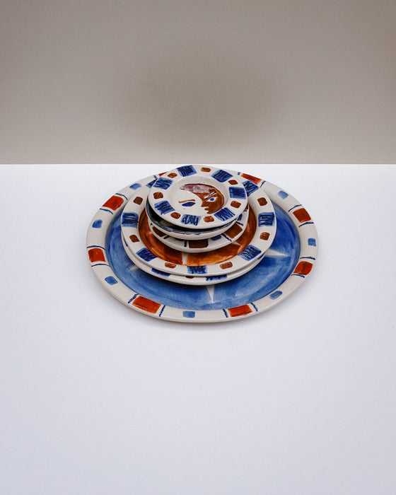 A stack of five nested ceramic plates with a hand-painted Majolica style design, featuring blue, terracotta orange, and white with small red accents. Hoodmor Studio | Fodder | Hope Collection