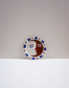 A round ceramic plate featuring a stylized face split into a brown moon and a white sun, with blue and brown decorative accents around the rim, set against a minimalist two-tone gray and white background. Hoodmor Studio | Fodder | Hope Collection