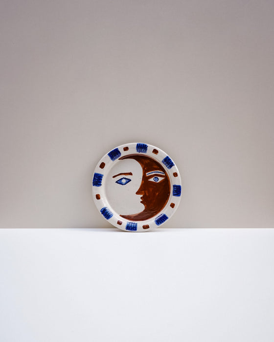 A round ceramic plate featuring a stylized face split into a brown moon and a white sun, with blue and brown decorative accents around the rim, set against a minimalist two-tone gray and white background. Hoodmor Studio | Fodder | Hope Collection