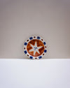 A ceramic plate with a rustic brown and white star design, featuring blue and brown accents on the rim, displayed on a white surface against a neutral gray background. Hoodmor Studio | Fodder | Hope Collection