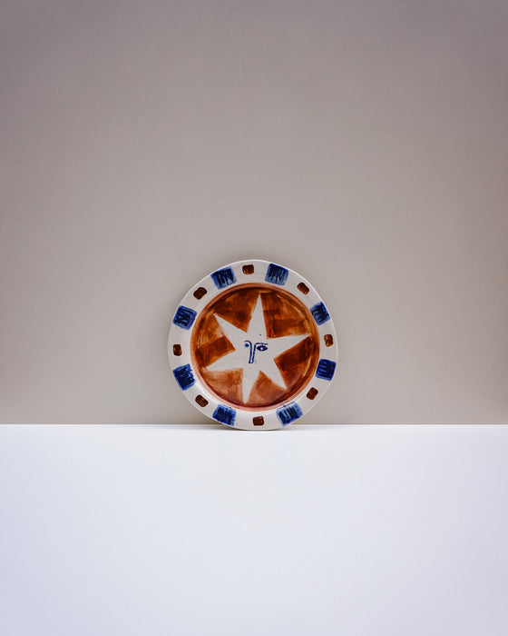 A ceramic plate with a rustic brown and white star design, featuring blue and brown accents on the rim, displayed on a white surface against a neutral gray background. Hoodmor Studio | Fodder | Hope Collection