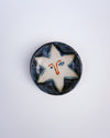 A round handmade ceramic plate featuring a stylized star design with a face, set against a plain light gray background. Hoodmor Studio | Fodder | Hope Collection
