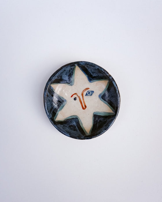 A round handmade ceramic plate featuring a stylized star design with a face, set against a plain light gray background. Hoodmor Studio | Fodder | Hope Collection