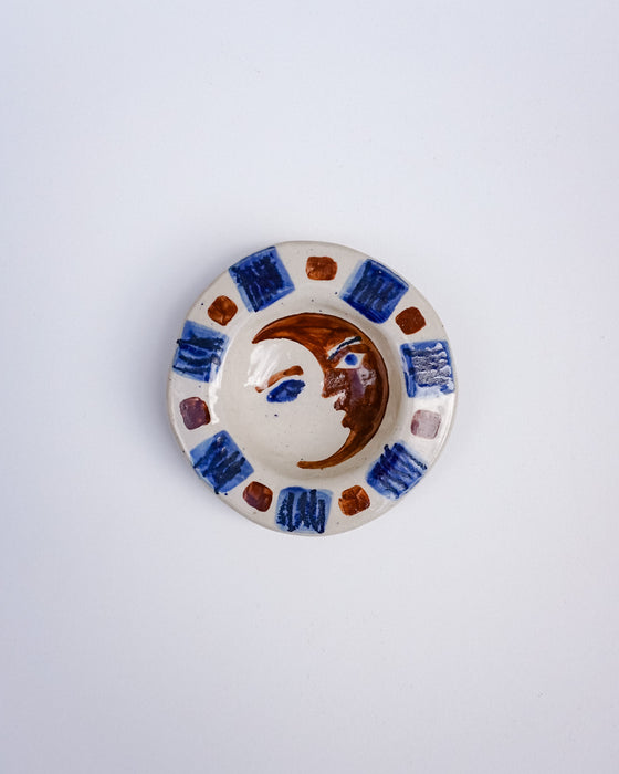 A small handmade ceramic dish with a hand-painted face in the center and a patterned border of blue, brown, and purple tiles on a white background. Hoodmor Studio | Fodder | Hope Collection