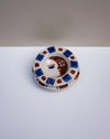 A handmade ceramic plate featuring a stylized face with blue and brown details, surrounded by a pattern of blue and brown squares on a white background. Hoodmor Studio | Fodder | Hope Collection