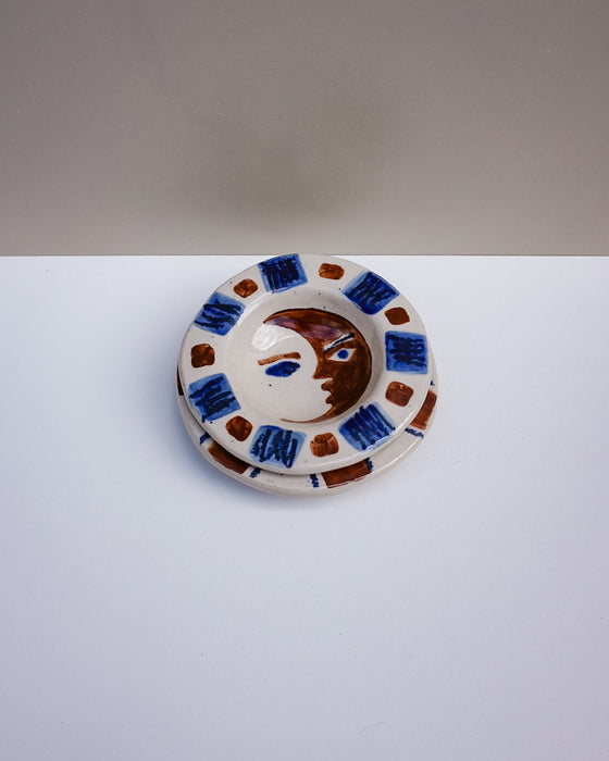 A handmade ceramic plate featuring a stylized face with blue and brown details, surrounded by a pattern of blue and brown squares on a white background. Hoodmor Studio | Fodder | Hope Collection
