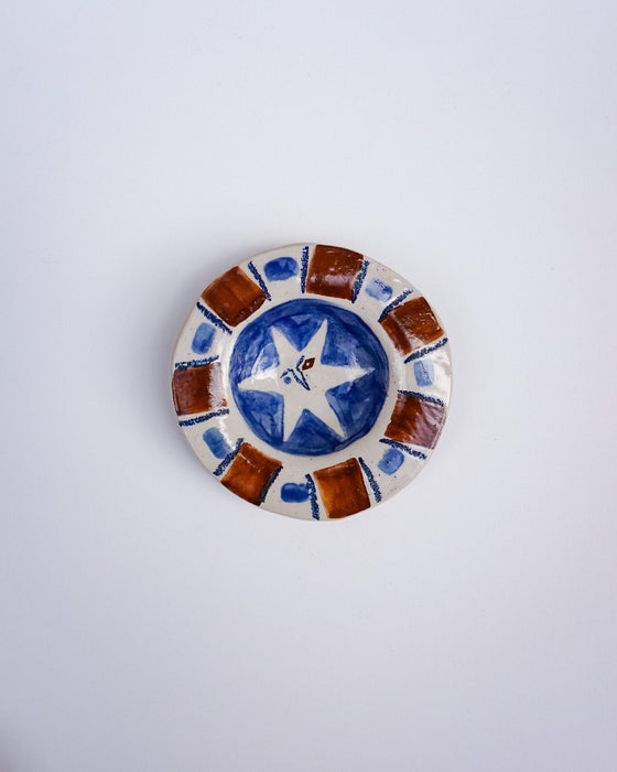 A small round ceramic dish with a hand-painted design featuring a blue and white star in the center and brown and white geometric border patterns, set against a plain light gray background. Hoodmor Studio | Fodder | Hope Collection