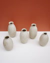 Five small speckled ceramic vases in a light warm gray-beige glaze, arranged on a white surface against a terracotta-colored background. KJA Studio Ceramics | Hope Collection | Shop Fodder
