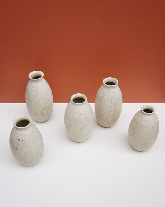 Five small speckled ceramic vases in a light warm gray-beige glaze, arranged on a white surface against a terracotta-colored background. KJA Studio Ceramics | Hope Collection | Shop Fodder