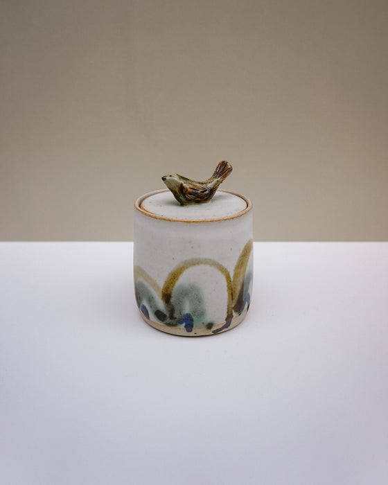 A small handmade ceramic lidded jar with a bird-shaped lid topper, featuring a white glaze with gold, green, and blue accents, set against a neutral beige and white background.  Liz Vidal Ceramics | Fodder | Hope Collection