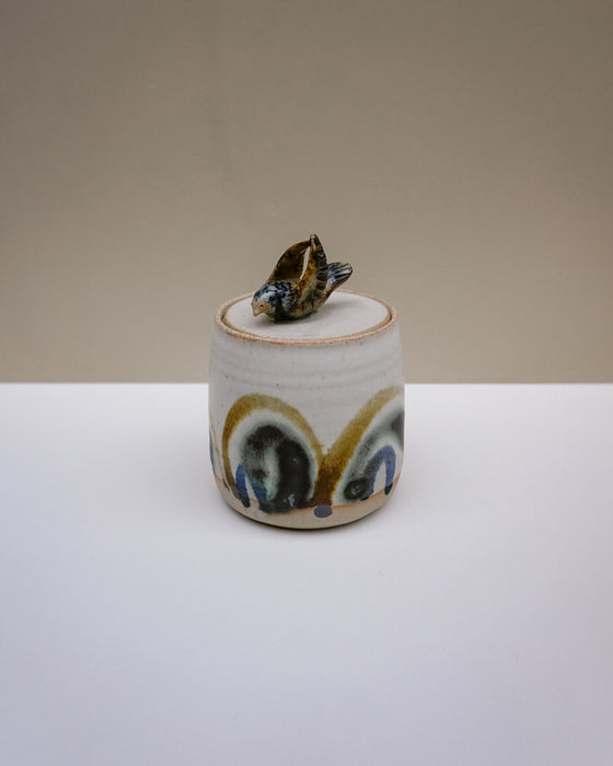 A small handmade ceramic lidded container with a lid topped by a small seashell figurine. The ceramic body is off-white with a glazed finish and features abstract painted patterns in blue, gold, and brown.  Liz Vidal Ceramics | Fodder | Hope Collection
