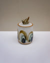 A small handmade ceramic lidded container with a bird figurine on top, featuring a white base with blue, green, and brown painted arch motifs, set against a neutral beige and white background.  Liz Vidal Ceramics | Fodder | Hope Collection
