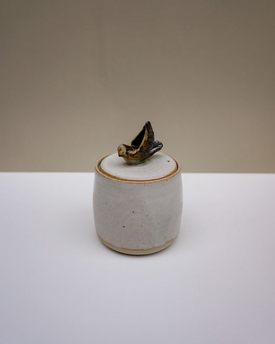A small handmade ceramic lidded container with a speckled off-white body and a dark brown to black organic-shaped lid topper, set against a neutral beige and white background.  Liz Vidal Ceramics | Fodder | Hope Collection