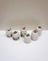 Six small cylindrical ceramic containers with lids, decorated with hand-painted patterns in green, black, and gold, arranged on a white surface against a neutral beige background.  Liz Vidal Ceramics | Fodder | Hope Collection