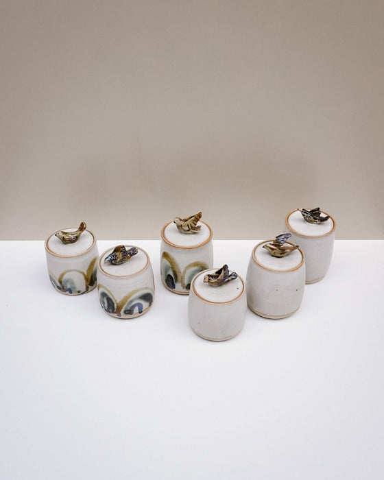Six small cylindrical ceramic containers with lids, decorated with hand-painted patterns in green, black, and gold, arranged on a white surface against a neutral beige background.  Liz Vidal Ceramics | Fodder | Hope Collection