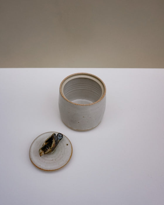 A handmade ceramic jar with its lid off, showing a small bird figurine inside. The jar has a light gray glaze with a rustic brown rim, and the lid is off to reveal the bird. Liz Vidal Ceramics | Fodder | Hope Collection